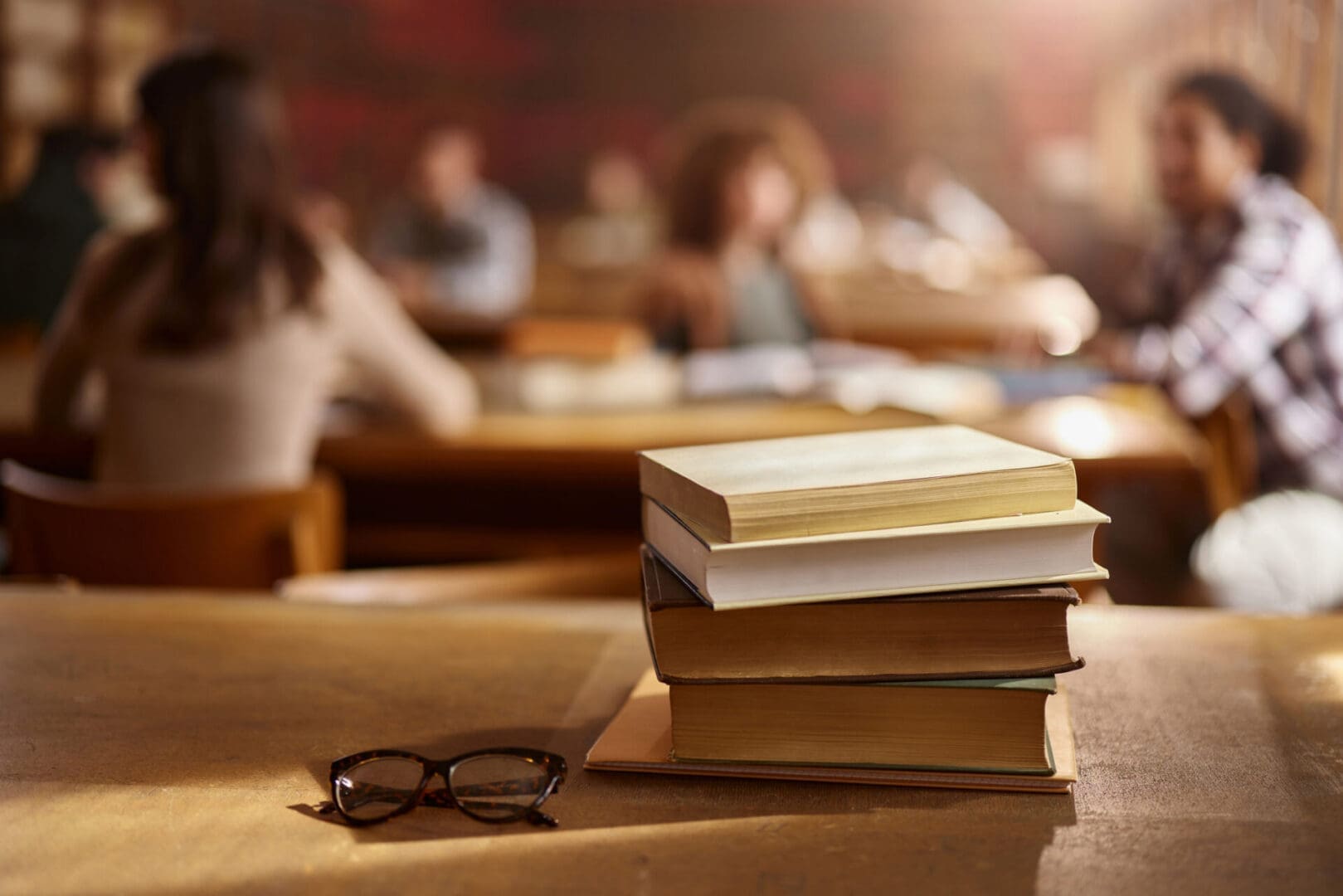 Close up of pile of books on a desk in library with people in the background.